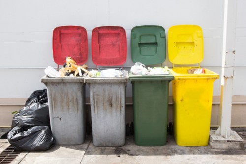 Four large commercial waste bins are positioned in a row outdoors, each with a different coloured lid: red, red, green, and yellow. The bins are made of hard plastic, with the lower bodies showing signs of weathering, and are filled with assorted rubbish, including paper, plastic bags, and miscellaneous waste materials. The red-lidded bins are on the left, with paper and plastic packaging visible over the edges, while the green bin in the middle appears to contain similar waste. The yellow-lidded bin on the far right is slightly larger and also filled with waste, with some items protruding from the top. To the left of the grey bins, there are two tightly packed black plastic rubbish bags resting against the side of the first bin, and the bins are set against a plain white wall with a drain grate on the ground nearby, indicating a typical outdoor waste collection area in a commercial or residential setting near Crawley. The scene is lit evenly, suggesting natural daylight, and the location subtly reflects the context of rubbish removal services provided by Commercial Waste Crawley in the local postcode area.