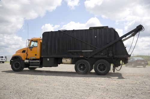 A large commercial waste collection vehicle with a yellow front cab and black rear body parked on a gravel surface, with a clear blue sky and scattered white clouds overhead. The vehicle has a robust, industrial design with a textured, ribbed metal waste container. The black rear section is fitted with an arm for emptying waste bins, extending diagonally upward and to the right, with a chain mechanism visible at the end. The vehicle's wheels are substantial and black, suitable for heavy-duty work, and a small white vehicle is visible parked in the background to the left, along with other vehicles more distant on the gravel lot. The surrounding environment appears to be an open, outdoor storage or waste management area, consistent with commercial waste removal operations near Crawley and the South East. Commercial Waste Crawley provides rubbish collection services, exemplified by this example of a waste collection truck used in local waste management workflows.