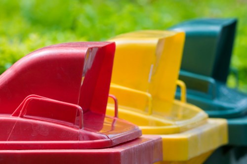 Close-up of a row of three plastic recycling bins placed outdoors on a grassy area, with a green background. The first bin on the left is red with a curved lid and a small handle, the middle bin is yellow with a similarly shaped lid and handle, and the third bin is teal with a matching lid. The bins are positioned side by side, slightly angled, indicating they are ready for waste collection or recycling disposal. The surface of the bins appears smooth and slightly reflective, with the bright colours contrasting against the natural greenery, emphasizing their purpose related to rubbish or waste management services in Crawley. This setup suggests a typical scene for waste collection points outside commercial or residential premises managed by companies like Commercial Waste Crawley, supporting effective rubbish removal and recycling efforts in the local area.