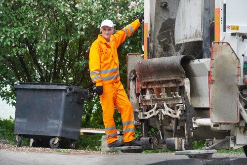 A waste collection worker dressed in a bright orange high-visibility suit with reflective striped bands, standing next to a large industrial rubbish compactor on a paved street. The worker is holding the handle of a black wheeled waste bin positioned to the left of the compaction machine, which is partially open revealing a collection of mixed waste materials inside. Behind the worker, there is dense green foliage and trees, indicating a suburban or semi-rural environment in the UK, possibly near Crawley. The compactor features a metallic grey finish with textured surfaces, and the surrounding pavement appears clean and well-maintained. The scene is lit by natural daylight, suggesting daytime, and the image visually supports professional rubbish and waste management services such as those provided by Commercial Waste Crawley, with a local context implied by the scene's setting on a residential or commercial street. The worker’s focused stance and the arrangement of the waste bin and machinery reflect standard practices for refuse collection and rubbish removal in the area.