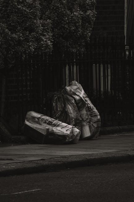 A scene of three large black plastic garbage bags, tightly packed and leaning against a black wooden fence on a street curb. The bags appear to contain household or garden waste, with some parts of th