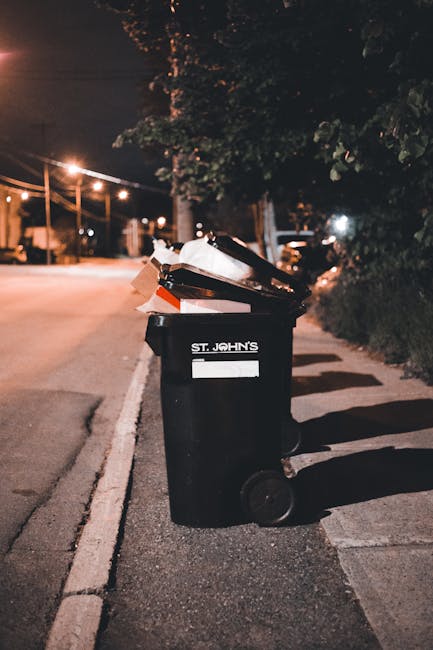 A scene of three large black plastic garbage bags, tightly packed and leaning against a black wooden fence on a street curb. The bags appear to contain household or garden waste, with some parts of th