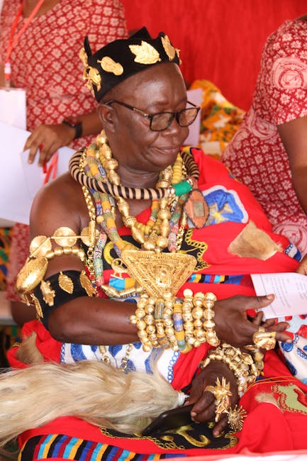 A woman dressed in traditional Maasai attire seated outdoors, wearing an elaborate headdress decorated with gold, vibrant beads, and intricate jewelry, including multiple necklaces, bracelets, and arm
