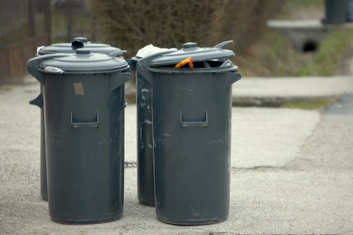 The image depicts three large, dark grey plastic wheelie bins positioned on a paved driveway or pavement, likely situated in a residential or commercial area in Crawley. The bins are cylindrical with smooth finishes, fitted with flat, matching lids. The leftmost bin has its lid securely closed, while the middle bin's lid is slightly open with visible trash bags or loose waste inside, including an orange item protruding from the opening. The rightmost bin appears to be closed as well. They are placed closely together with some signs of wear and scratches on the surfaces. In the background, there are trees and a grey stone or concrete bridge, suggesting an outdoor setting during daylight hours with overcast weather. These bins are typical of waste collection services, such as those provided by Commercial Waste Crawley, which manage rubbish removal for local households and businesses within the Crawley area, including nearby postcode districts. The scene emphasizes the essential aspect of waste management in maintaining cleanliness and environmental standards.